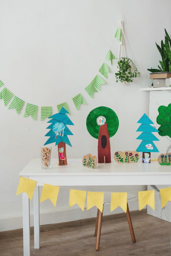 White table with handmade colorful trees placed against white walls in light room with garlands of paper flags devoted for children event